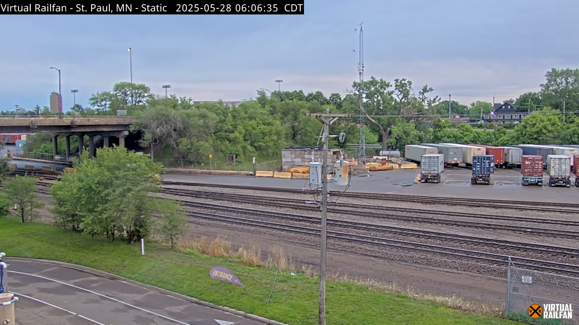 A train yard with multiple sets of railway tracks, several shipping containers, and a road overpass under an overcast sky.