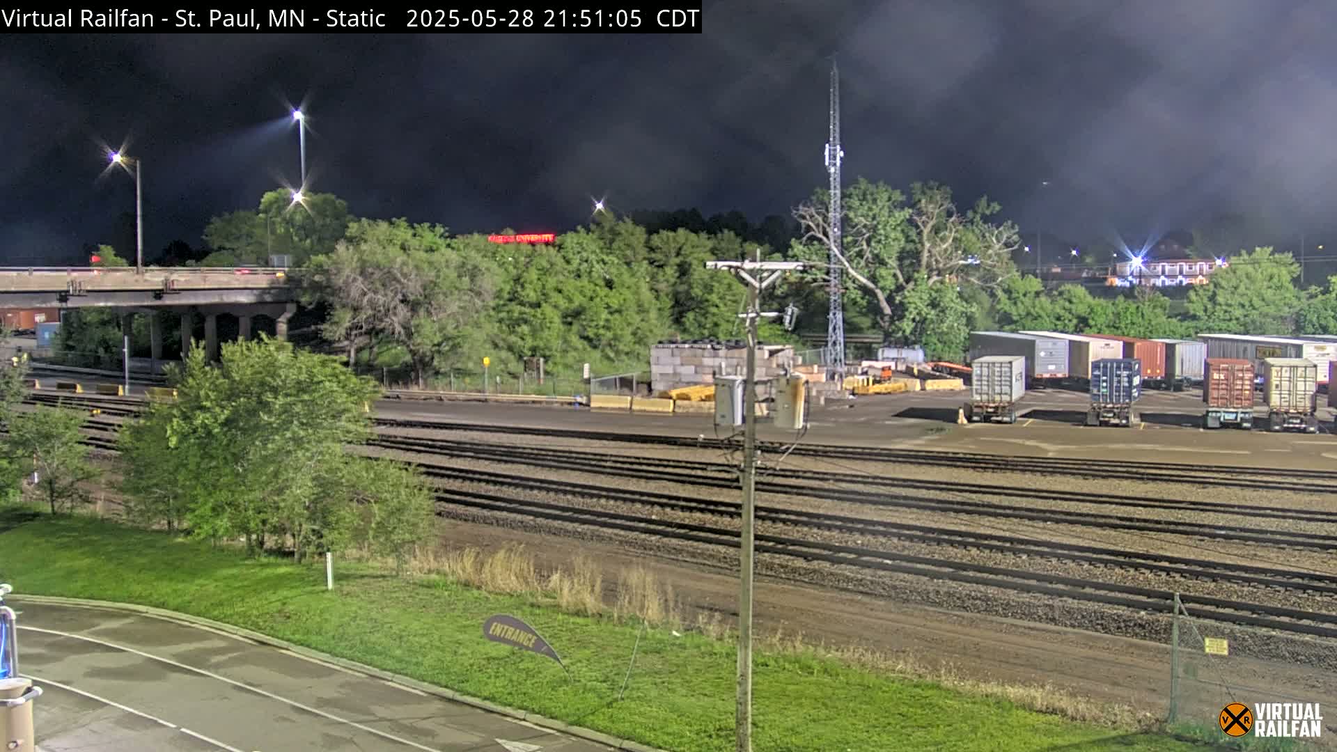 A nighttime view of a railway yard with several freight containers, a road and bridge, and lush green vegetation under a partly cloudy sky.