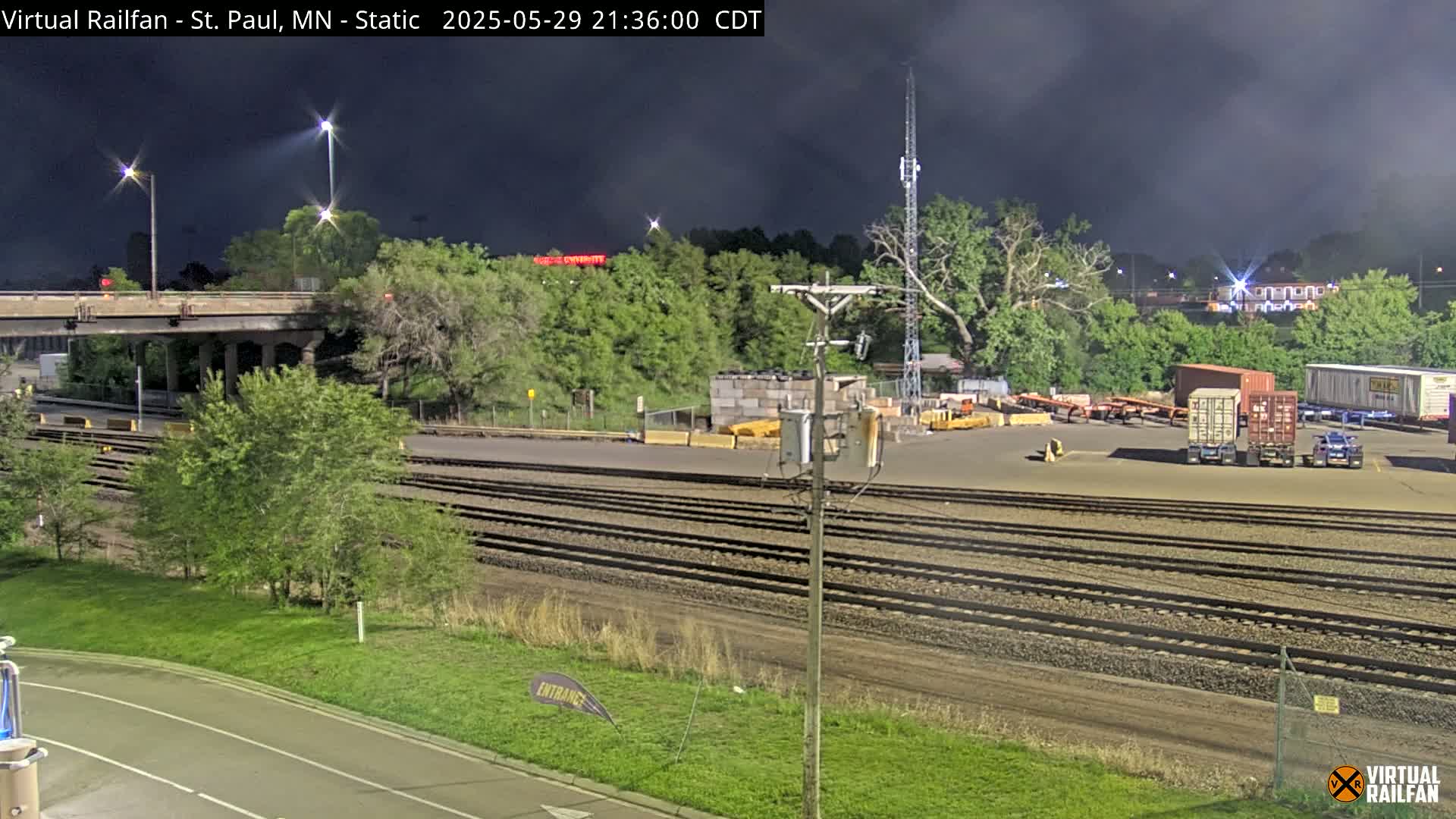 Under a dark, cloudy night sky, a railroad yard with several train cars and tracks is visible near a highway overpass, surrounded by green vegetation.