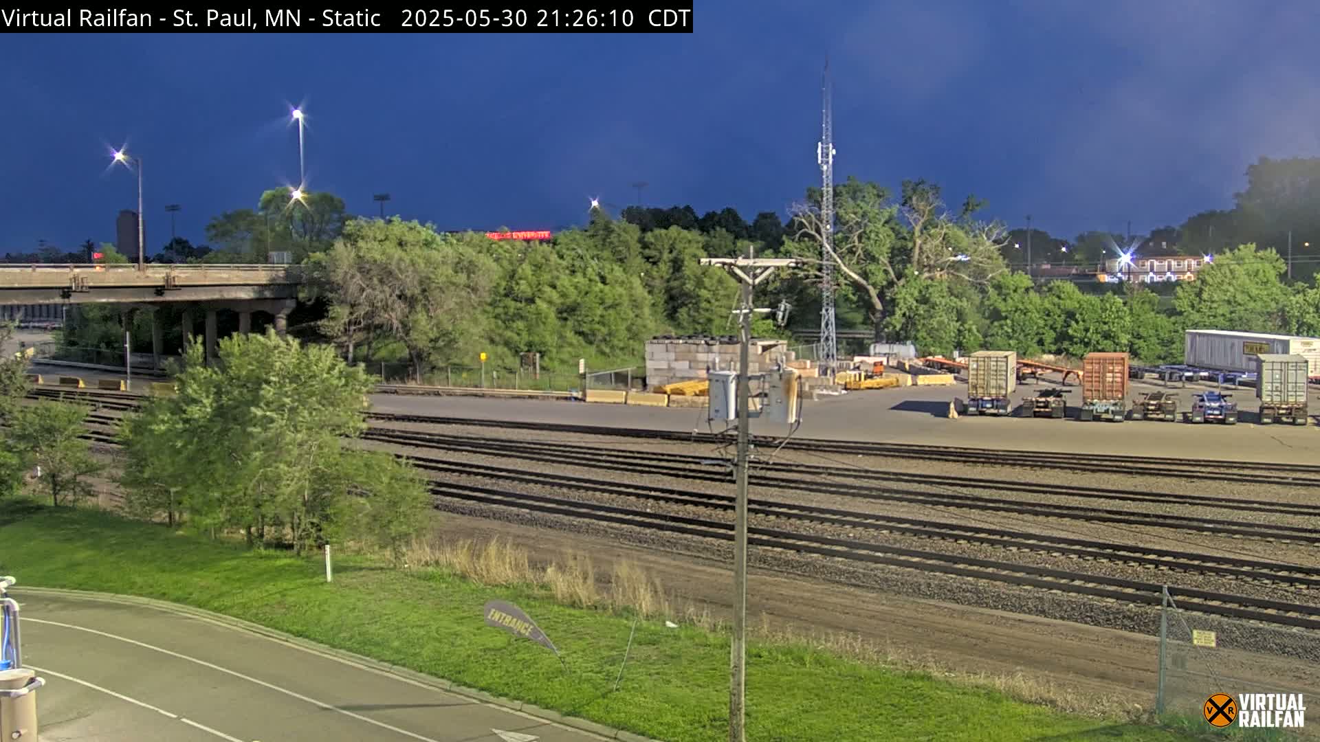 A twilight view of a rail yard with several shipping containers, train tracks, and a roadway under a bridge, surrounded by trees.