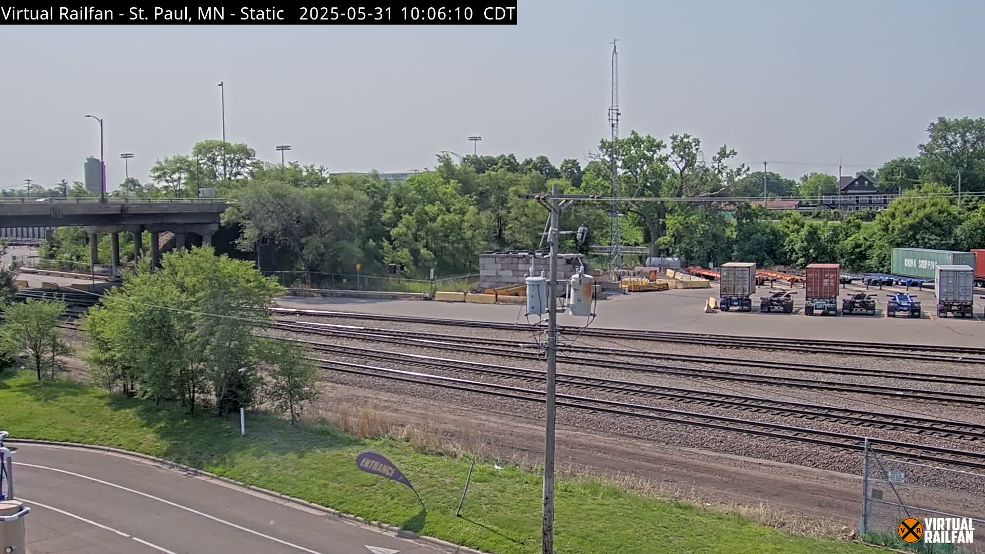 Under a clear sky, several freight containers sit in a lot adjacent to a section of train tracks that runs parallel to a road and under a bridge.
