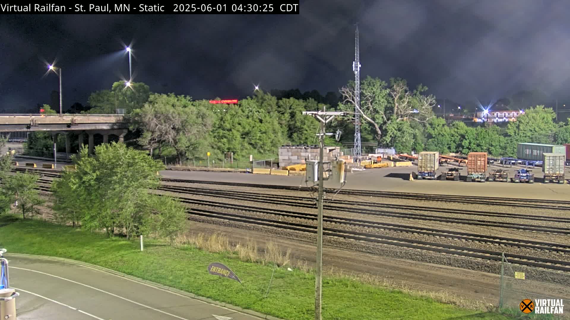 A nighttime view of a railroad yard with several freight containers, train tracks, and a roadway under a partly cloudy sky.