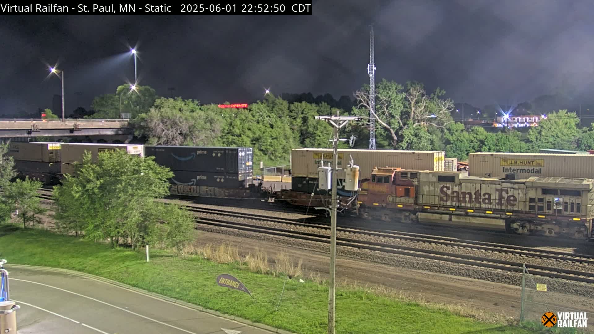 At night, under a dark, cloudy sky, a freight train with several containers, including some marked with Amazon logos, sits on railway tracks beside lush green vegetation.