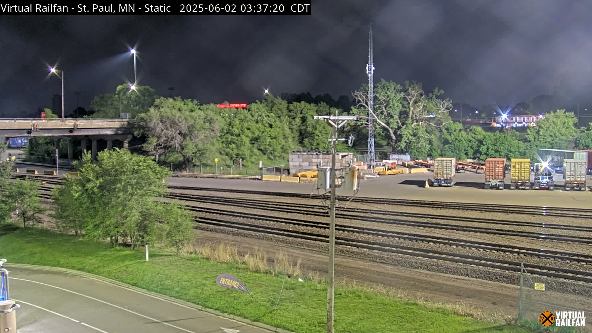 A nighttime view of a rail yard with several shipping containers, train tracks, and lush green vegetation under a partly cloudy sky.