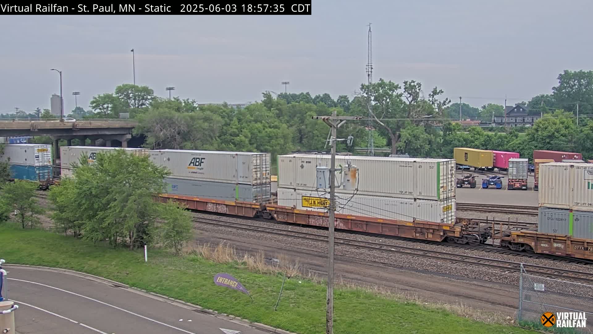 A train carrying multiple shipping containers sits on railroad tracks under an overcast sky, next to a road and green vegetation.