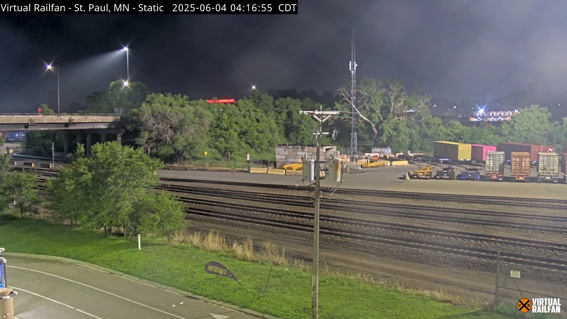 Under a dark, night sky, a train yard with several cargo containers sits next to a set of train tracks that run alongside a paved road and a grassy area with trees.