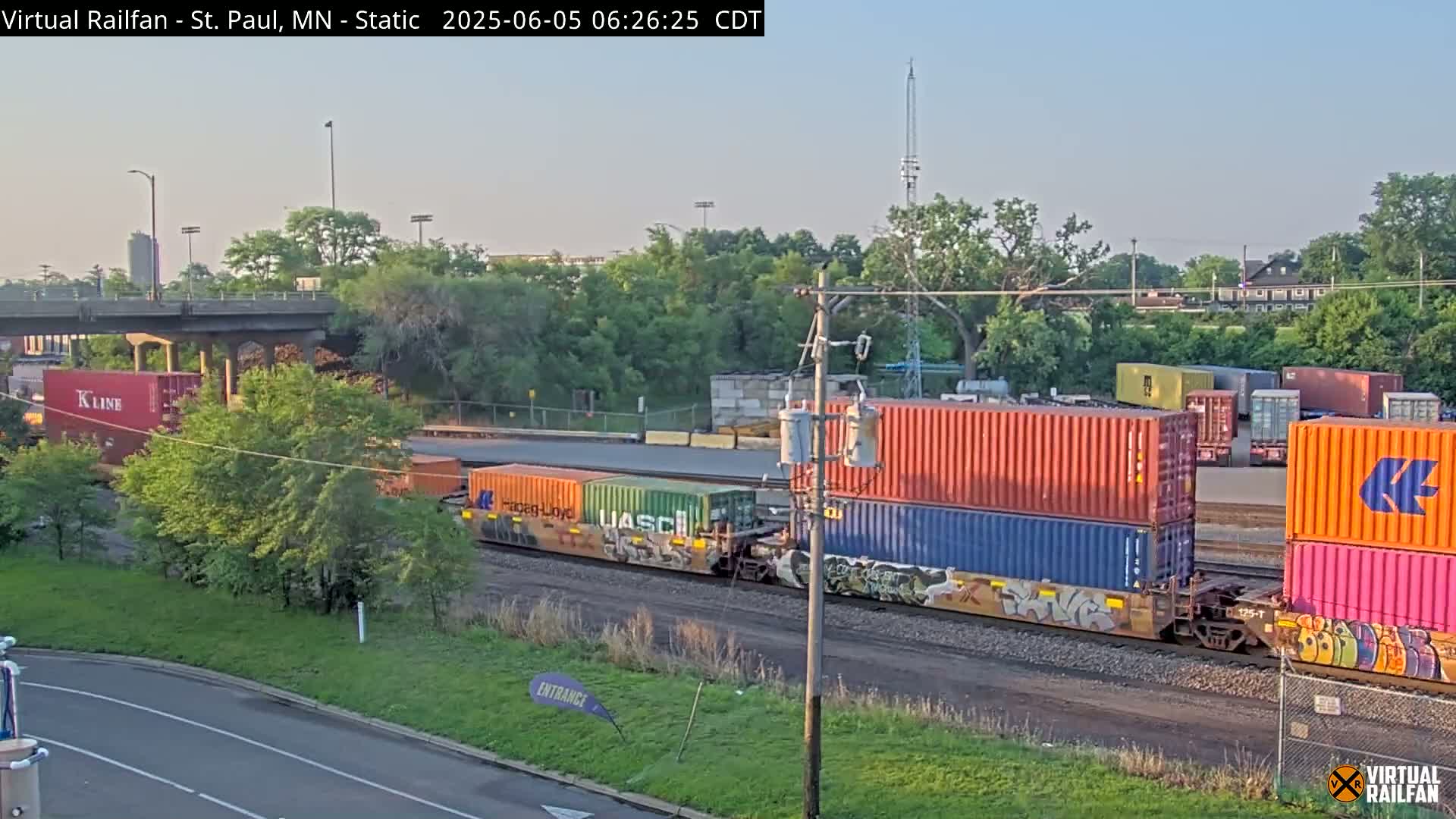 A freight train carrying colorful shipping containers is visible, partially obscured by trees and under a partly sunny sky.