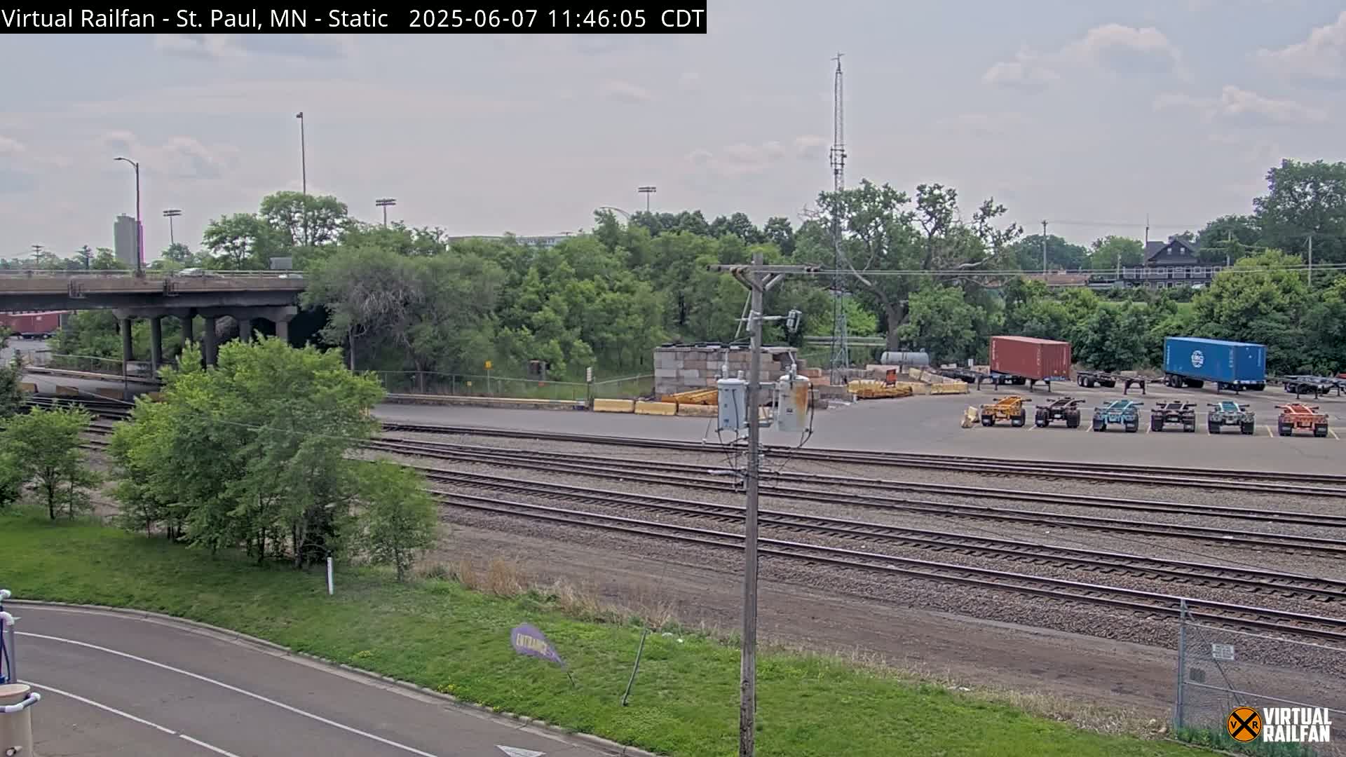 A view of railroad tracks and a yard with several shipping containers and trucks, under an overcast sky, next to a highway overpass.