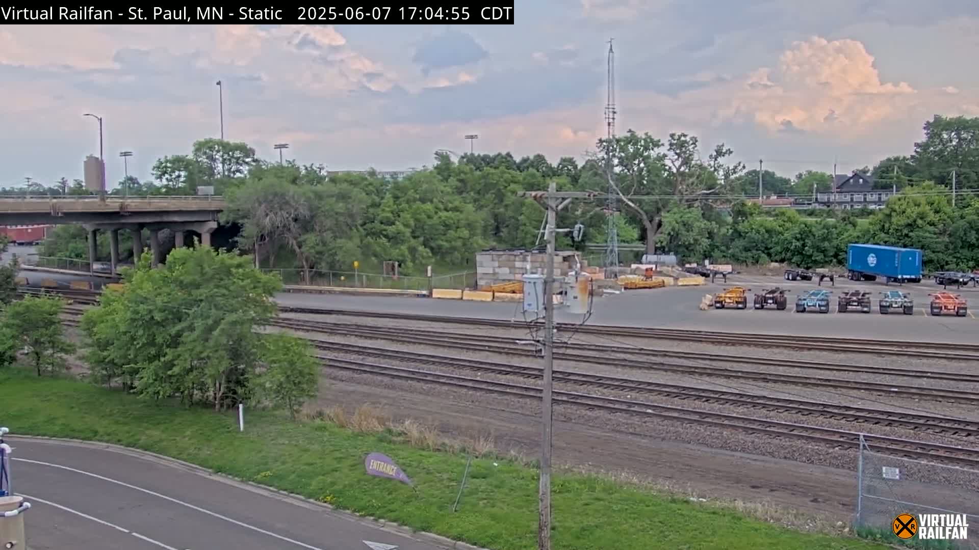 Railroad tracks and a paved area with several pieces of industrial equipment and a blue shipping container are visible under a partly cloudy sky.