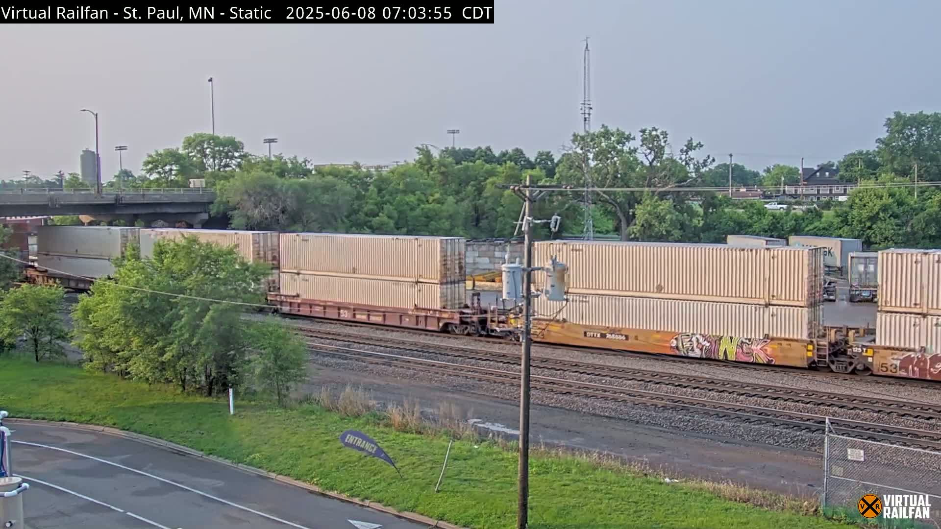 A long freight train carrying many stacked shipping containers moves along railroad tracks under a partly cloudy sky.