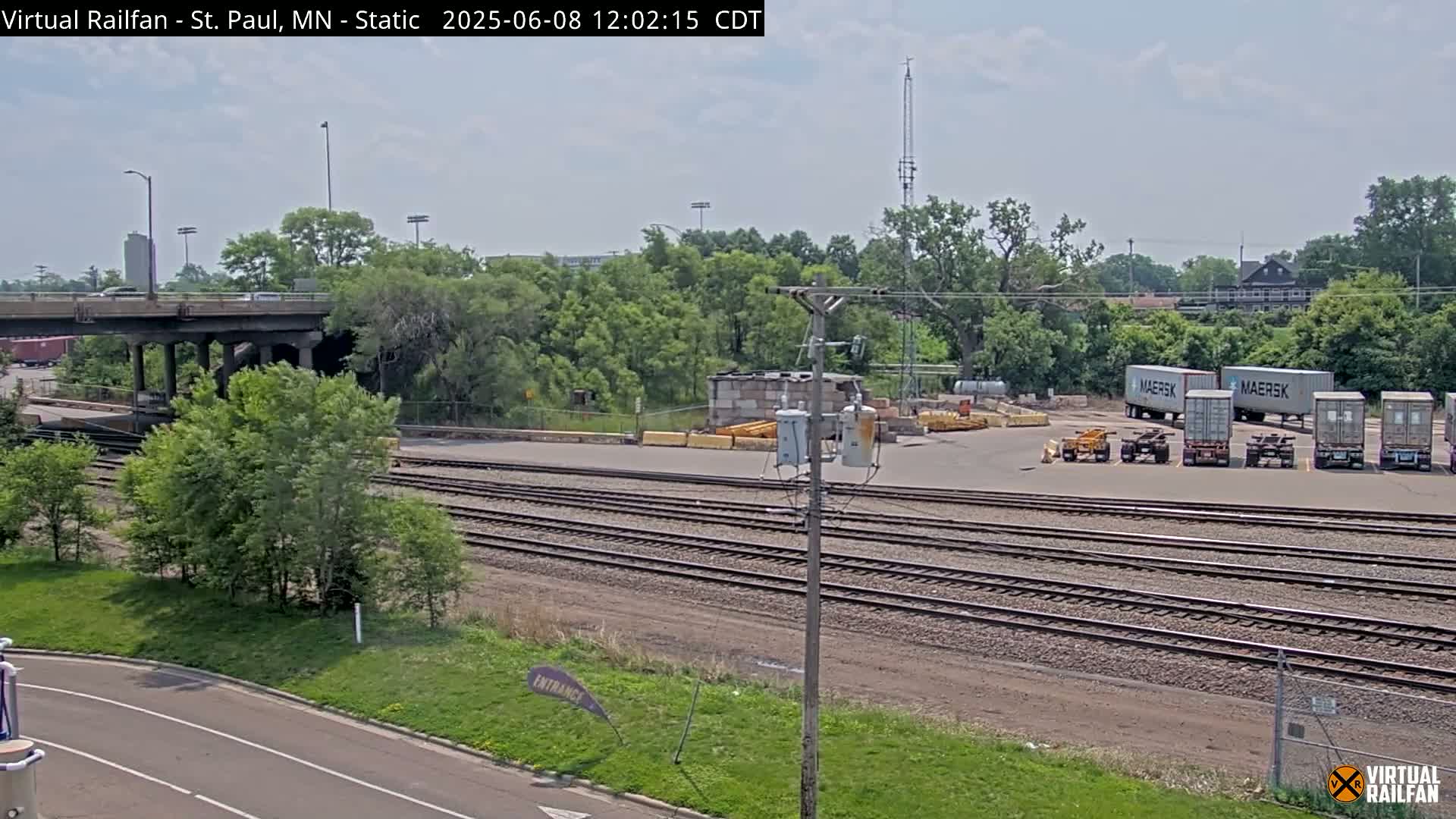 A view of railroad tracks and a paved area with several shipping containers under a partly cloudy sky.