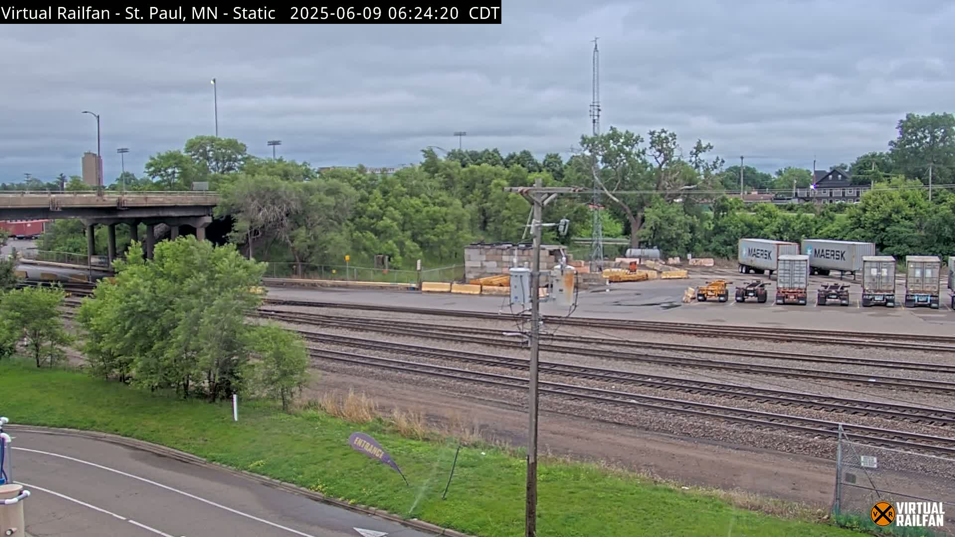 Under an overcast sky, a railroad yard with several shipping containers sits beside a road and a bridge over train tracks.