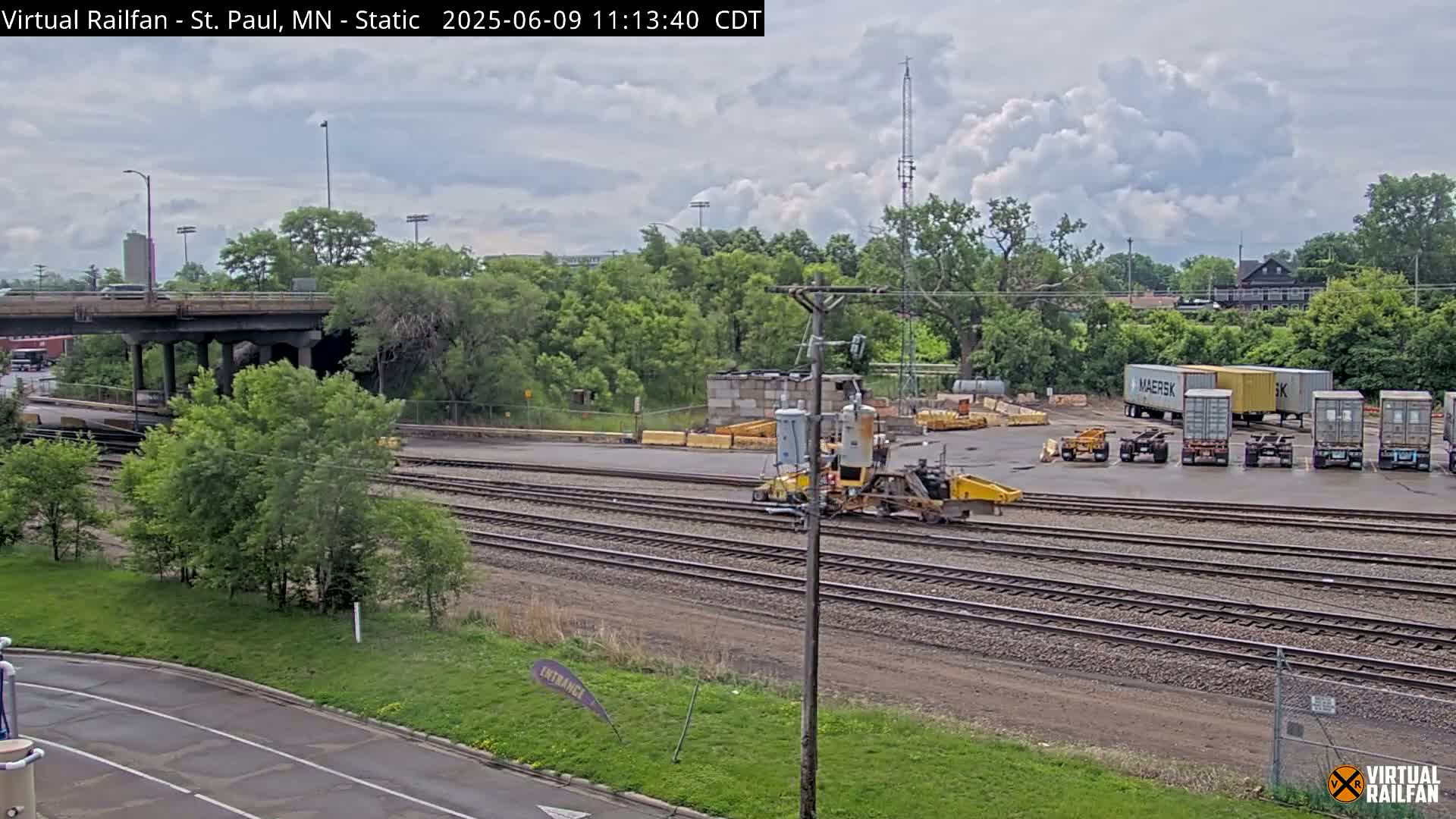 A partly cloudy day reveals a railroad yard with multiple train tracks, a yellow rail maintenance machine, several shipping containers, and a road overpass in the background.