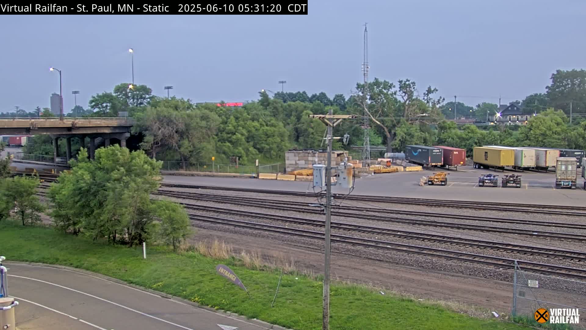 Under a hazy sky, a railroad yard with several shipping containers and train tracks is visible, alongside a road and an overpass.