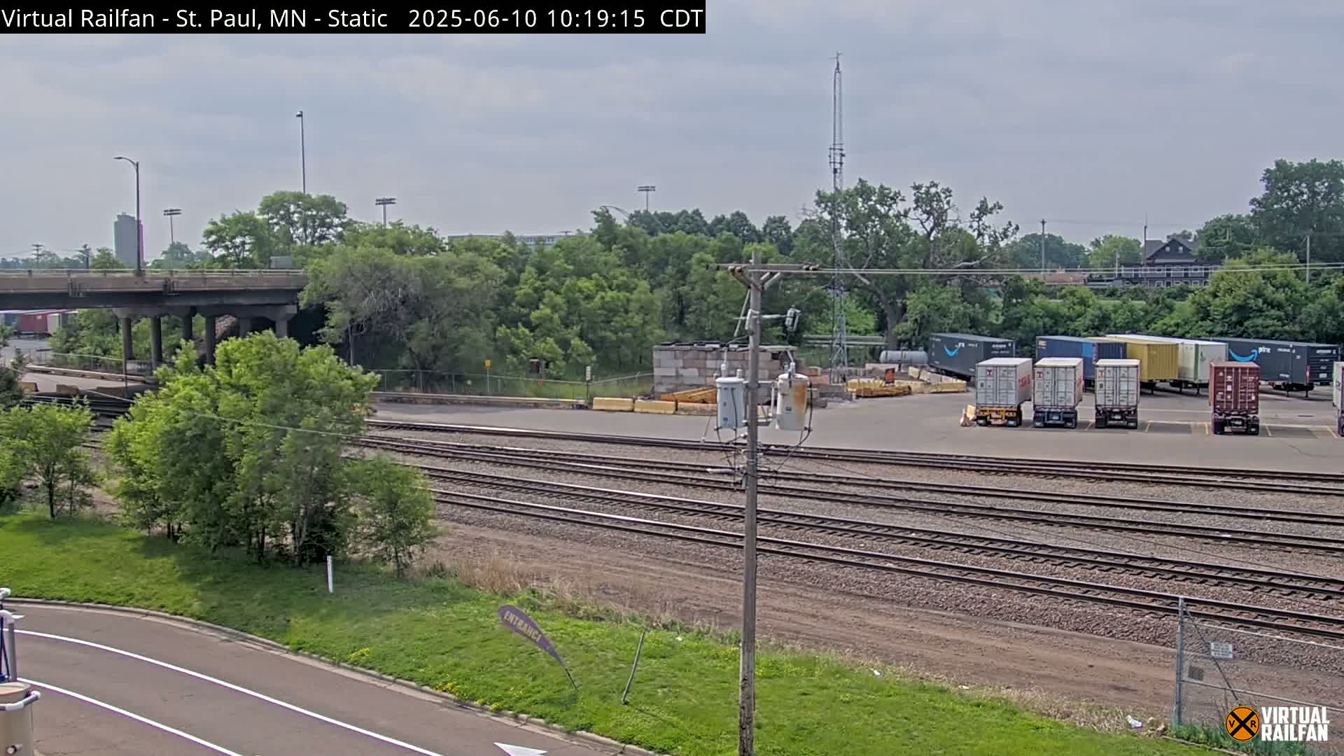 A railroad yard with several shipping containers, a road, a bridge, and lush green trees under a partly cloudy sky.