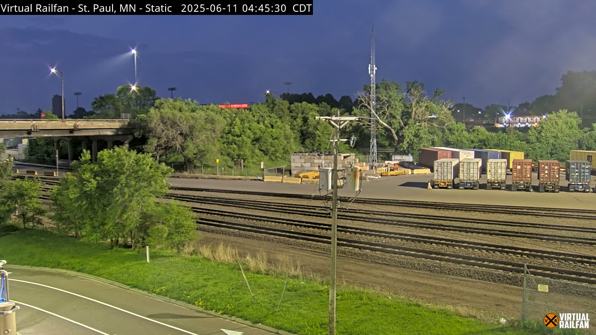Under a twilight sky, a railroad yard with multiple sets of tracks is shown, alongside a road, a bridge, and a line of shipping containers.