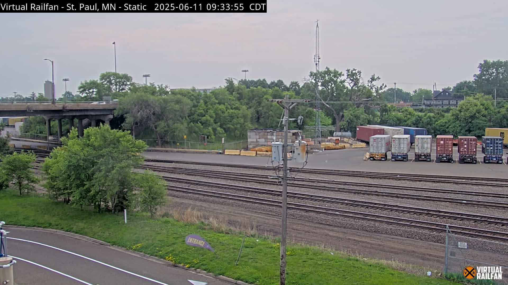 A cloudy day reveals a railroad yard with several shipping containers, train tracks, a road overpass, and lush green vegetation.