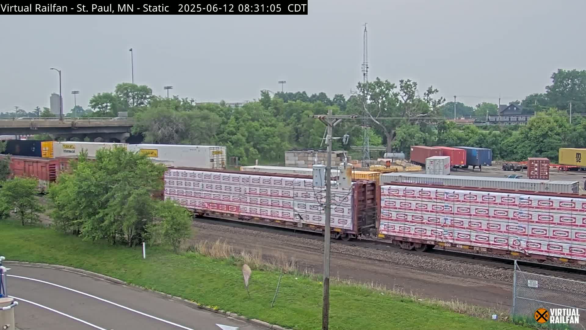 A long freight train carrying various cargo, including numerous bundles of sheathed lumber, is passing under an overpass on an overcast day.