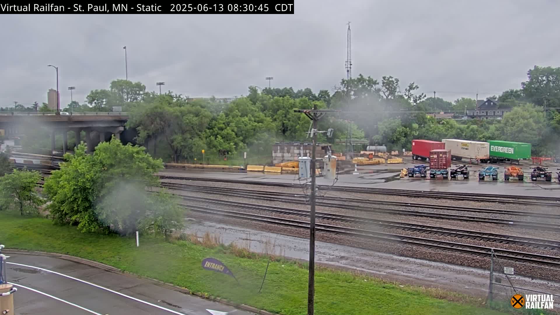 A rainy view of railroad tracks, a road, and a yard with shipping containers and other vehicles, surrounded by lush green trees and under a gray sky.