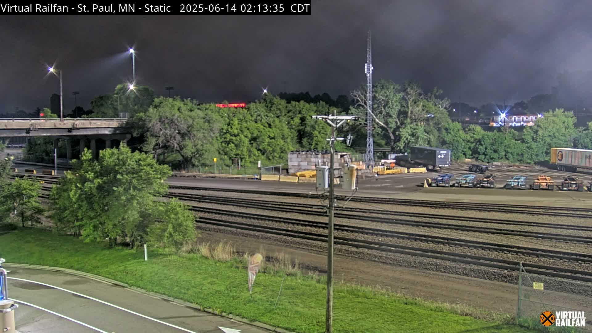 A nighttime view of a rail yard with several parked vehicles, surrounded by trees and a highway overpass under a dark, cloudy sky.