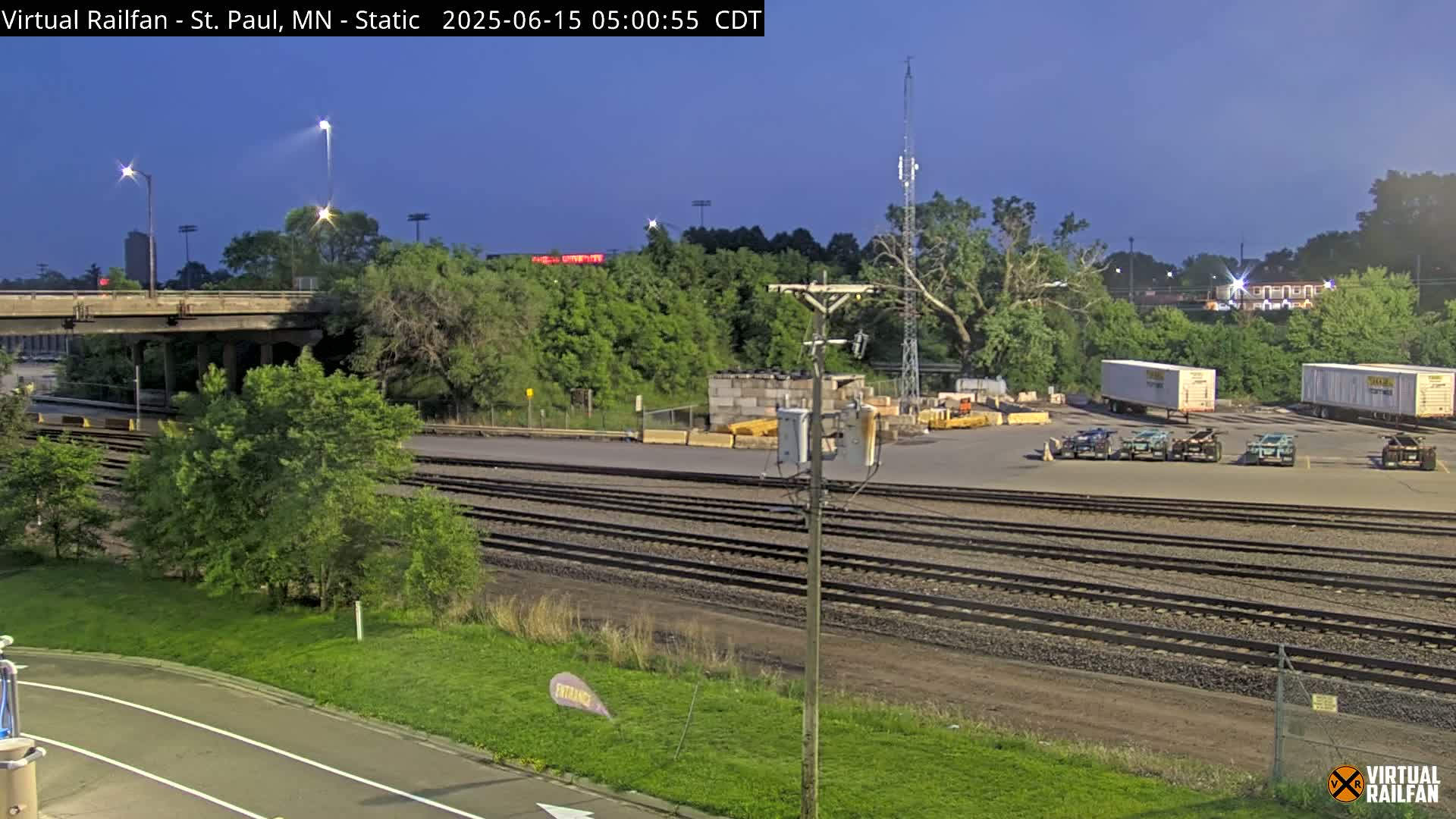 Under a twilight sky, a rail yard with several semi-trailers is visible, next to a paved road and lush green vegetation, alongside multiple train tracks.
