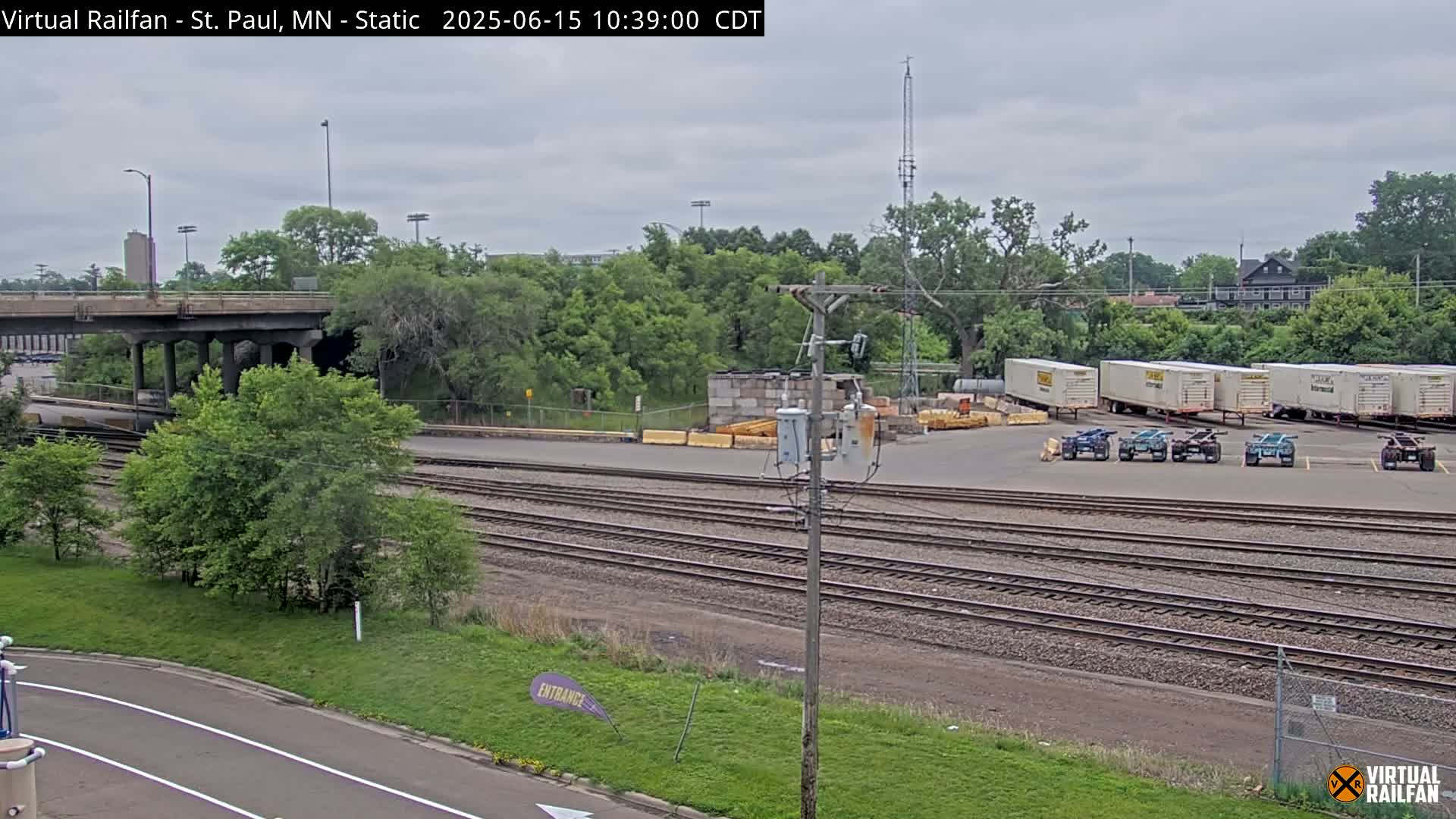 An overcast day reveals a railway yard with several freight trailers parked near a road and overpass, surrounded by lush green trees.