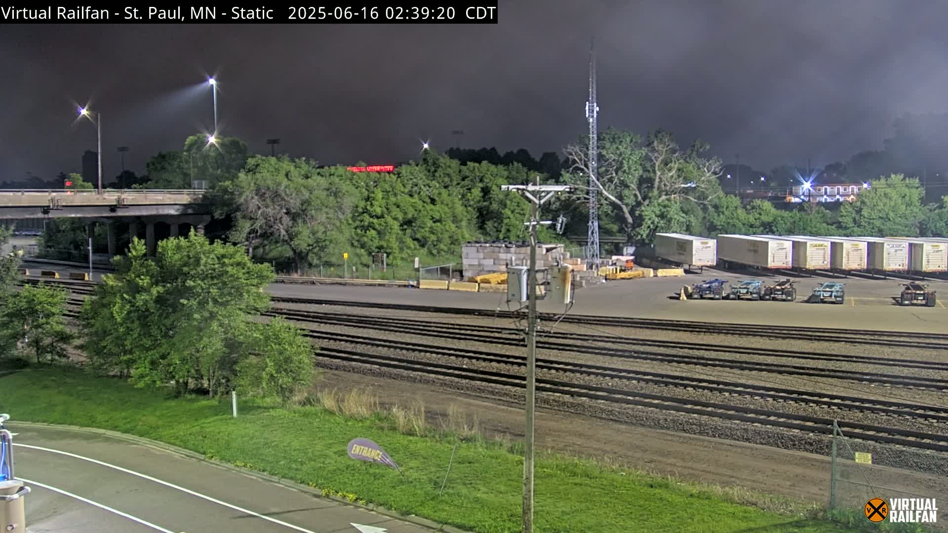 Under a dark, nighttime sky, a rail yard with numerous train cars and a roadway overpass is illuminated by artificial lights.