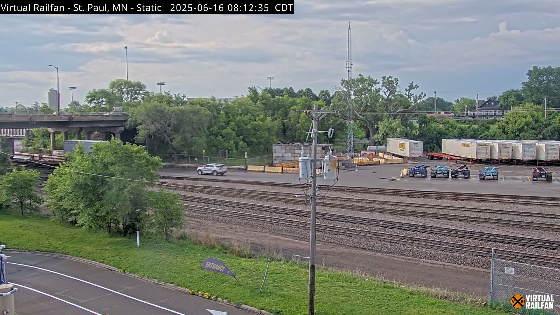 Under a partly cloudy sky, a railway yard with numerous freight containers and railcars is visible, alongside a road and some vegetation.