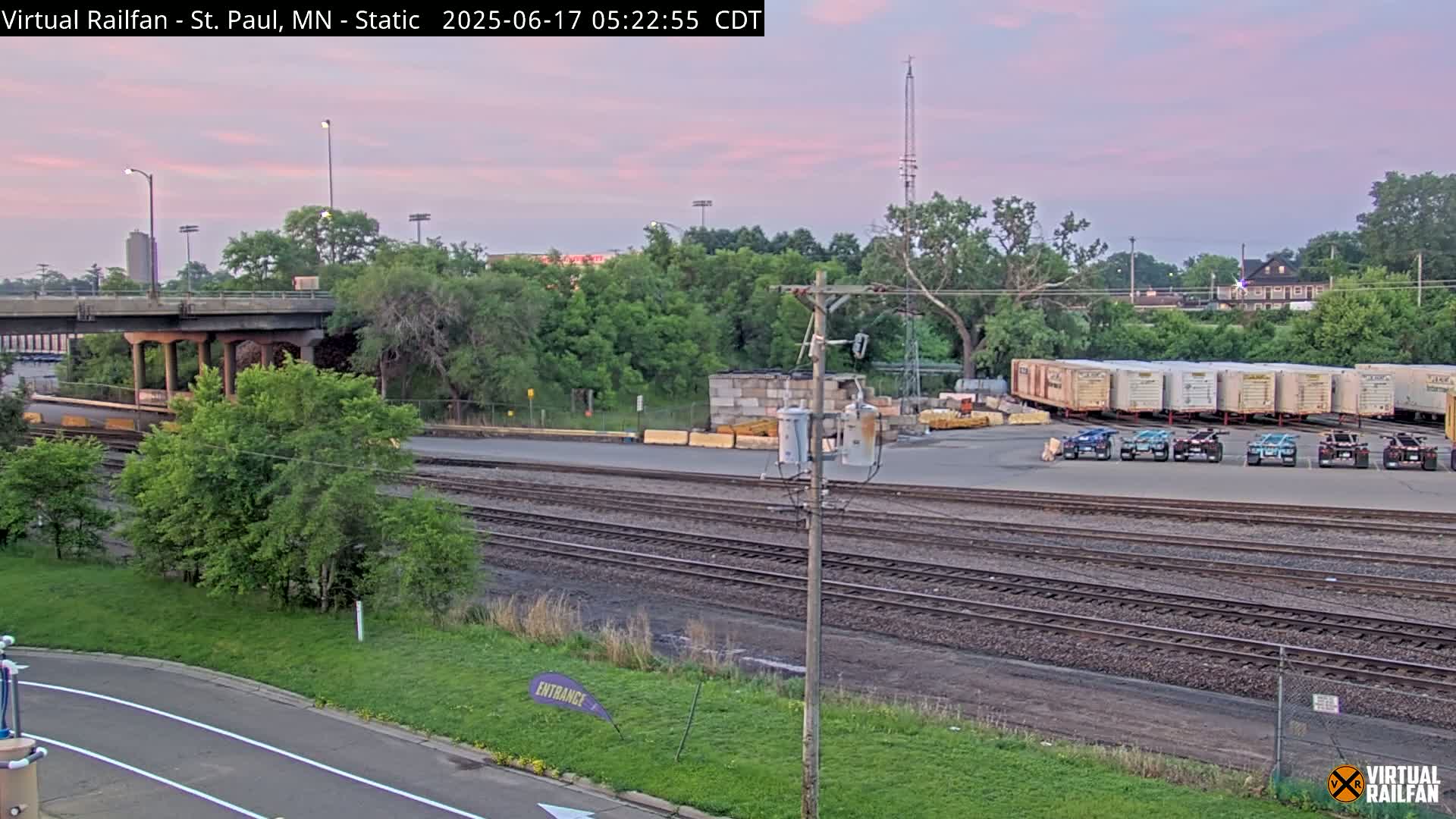A railway yard with several train cars and vehicles, under a pale pink twilight sky.