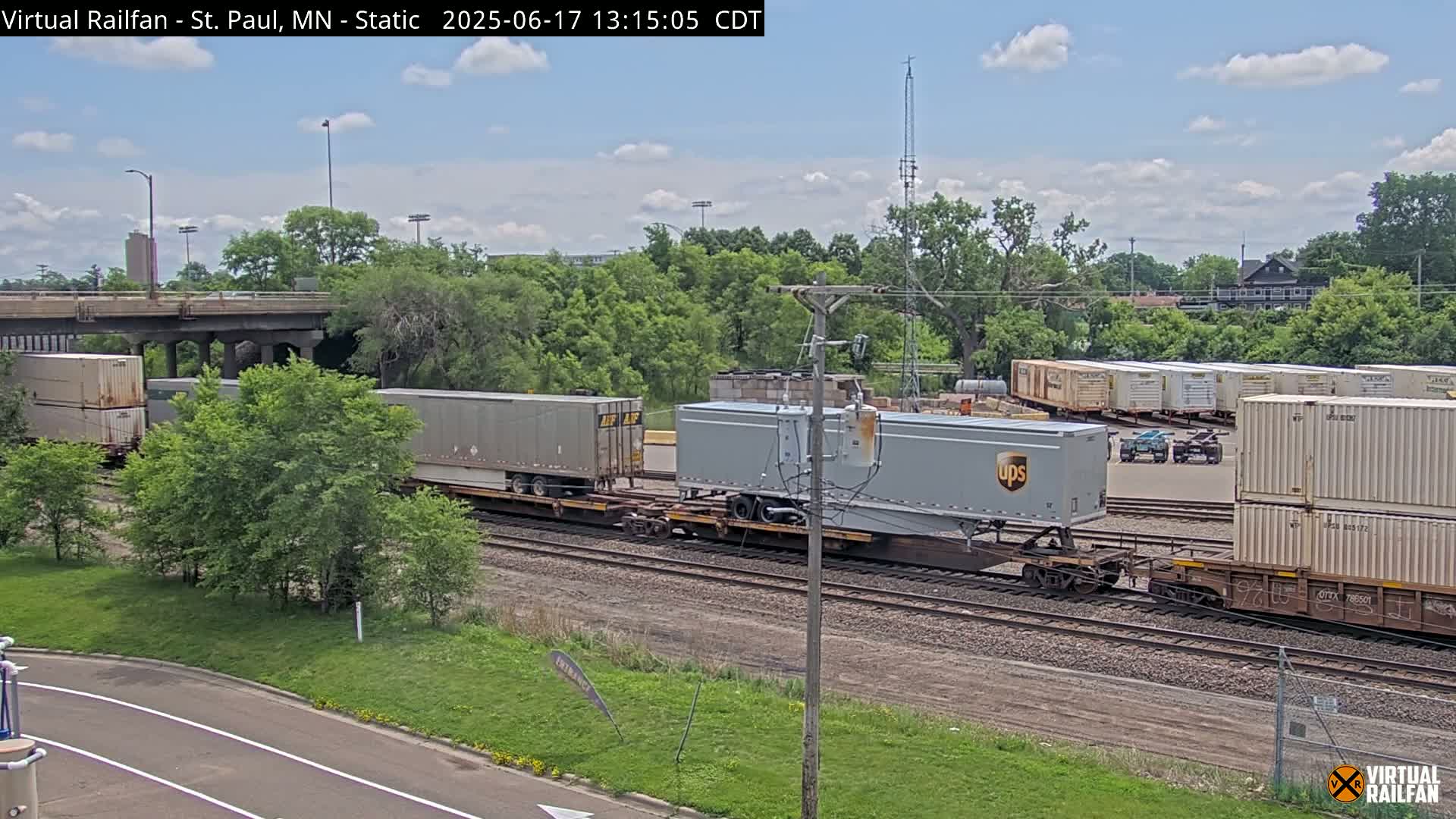 A freight train carrying shipping containers, including one marked UPS, passes under a bridge on a sunny day.
