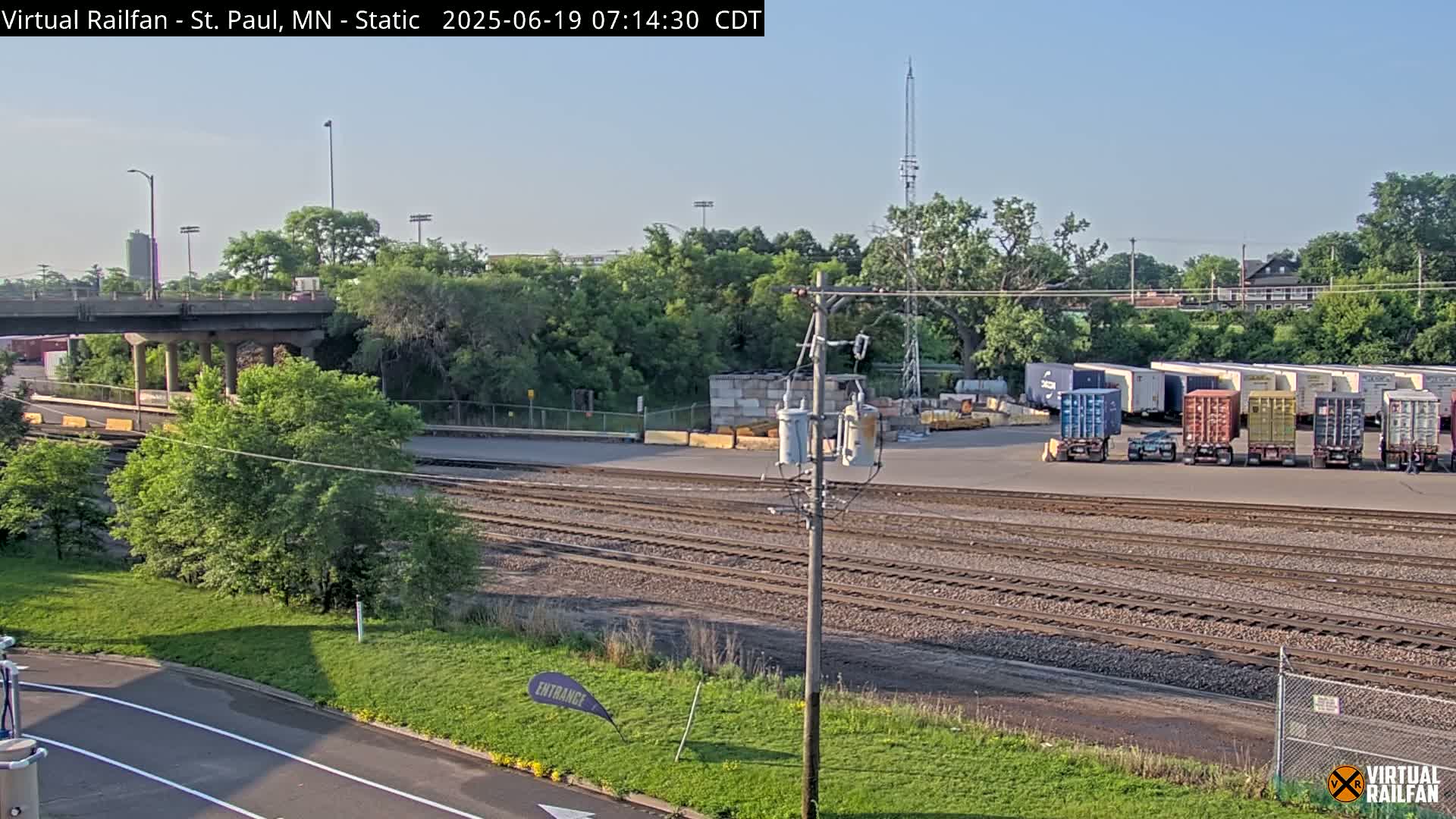 A sunny day reveals a railway yard with multiple shipping containers, train tracks, and a roadway overpass, surrounded by lush green vegetation.