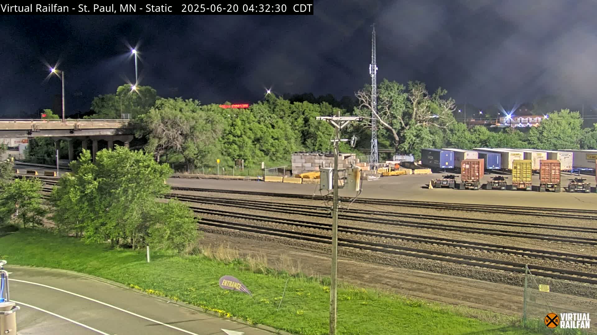Under a dark, cloudy night sky, a rail yard with several shipping containers sits beside a set of train tracks that runs under a highway overpass.