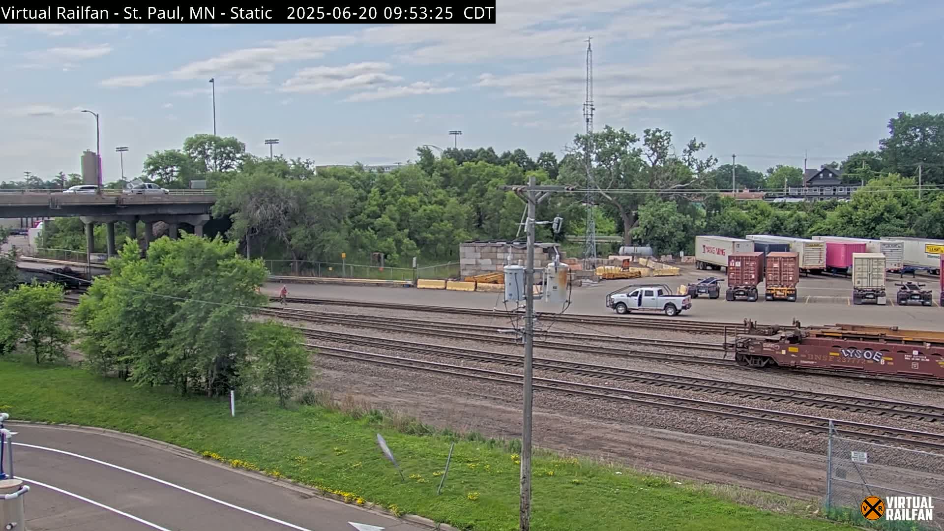A mostly sunny day reveals a view of railroad tracks, a highway overpass, several shipping containers, and some vehicles in a yard, with lush green vegetation surrounding the scene.