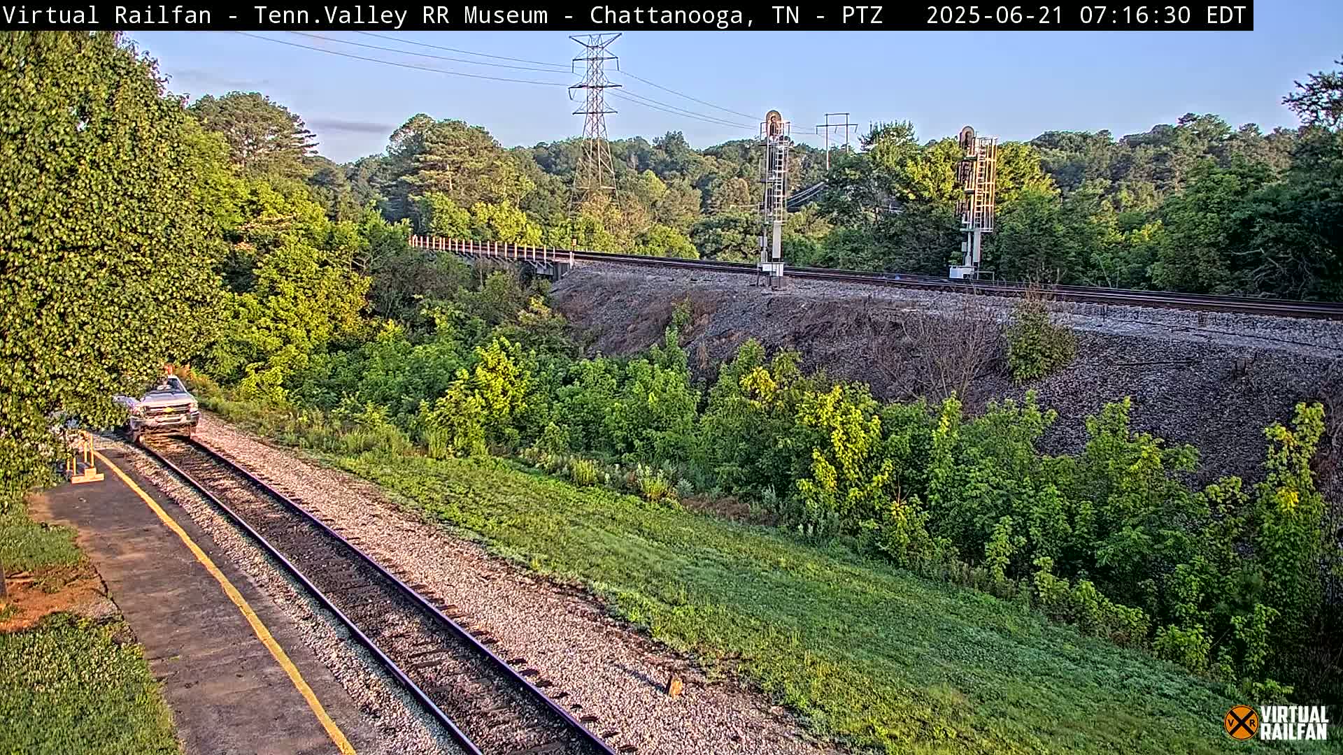 A light-colored truck is parked next to railroad tracks that run through a lush green landscape under a mostly clear sky.