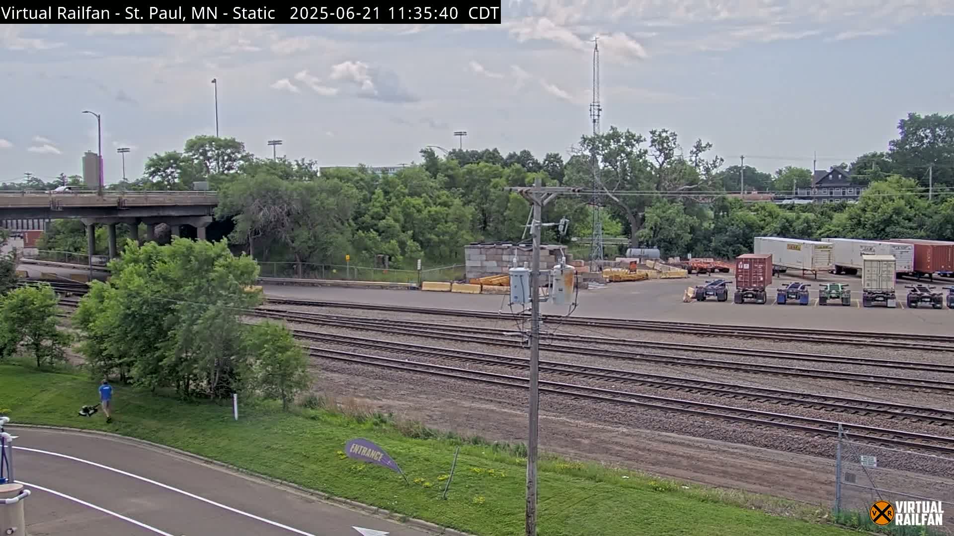 A partly cloudy day reveals a railway yard with several shipping containers, a road, and a person mowing a lawn near a bridge.