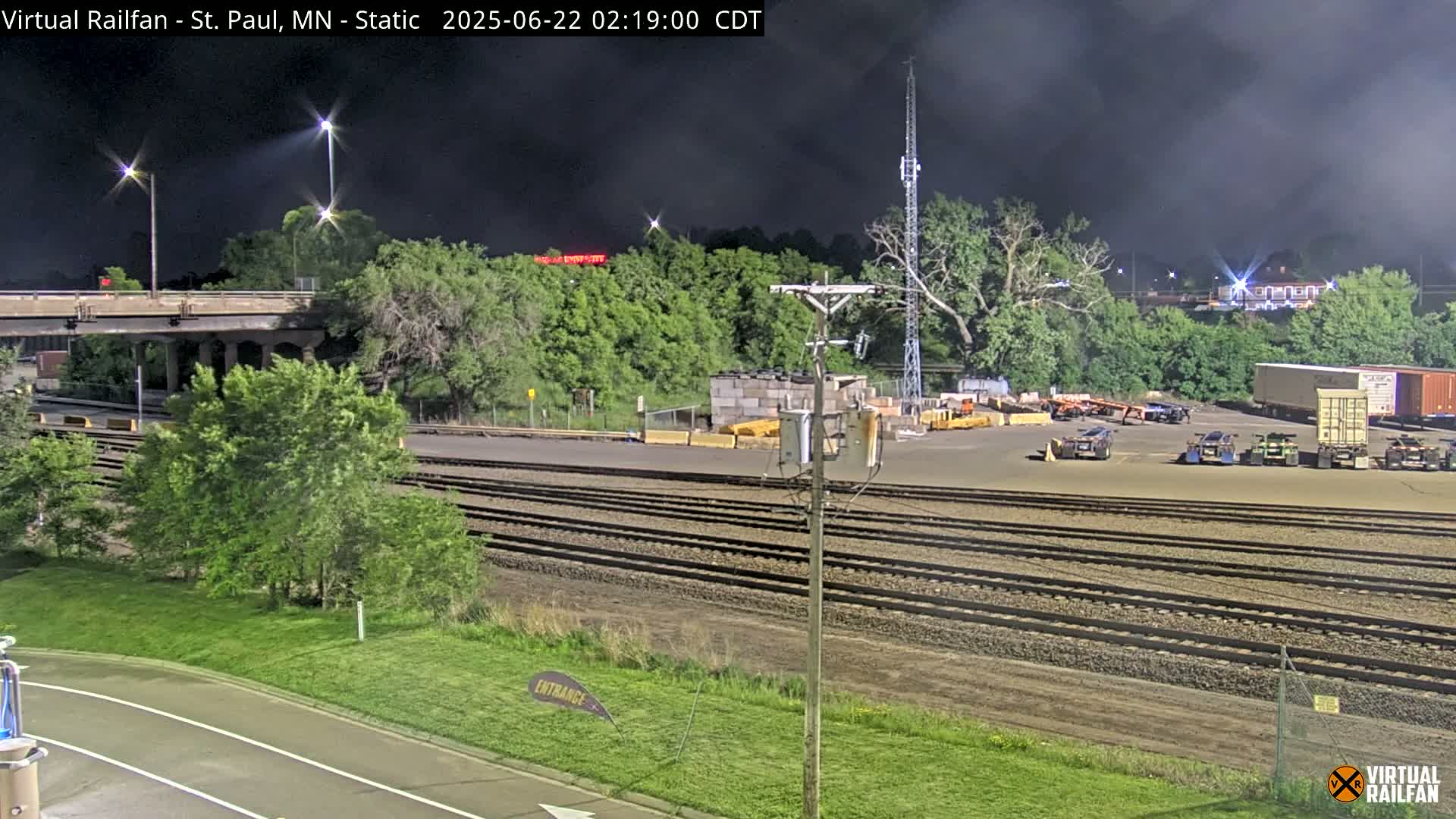 Under a dark, cloudy night sky, a railway yard with several parked vehicles and train tracks is visible next to a highway overpass and some green trees.
