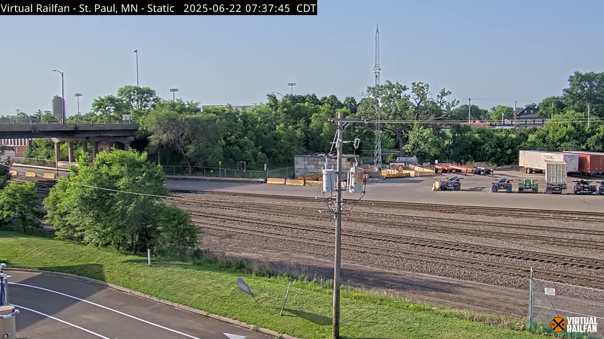 A sunny view of railroad tracks, a roadway, and a yard with several semi-trailers parked next to a lush green area, under a clear sky.