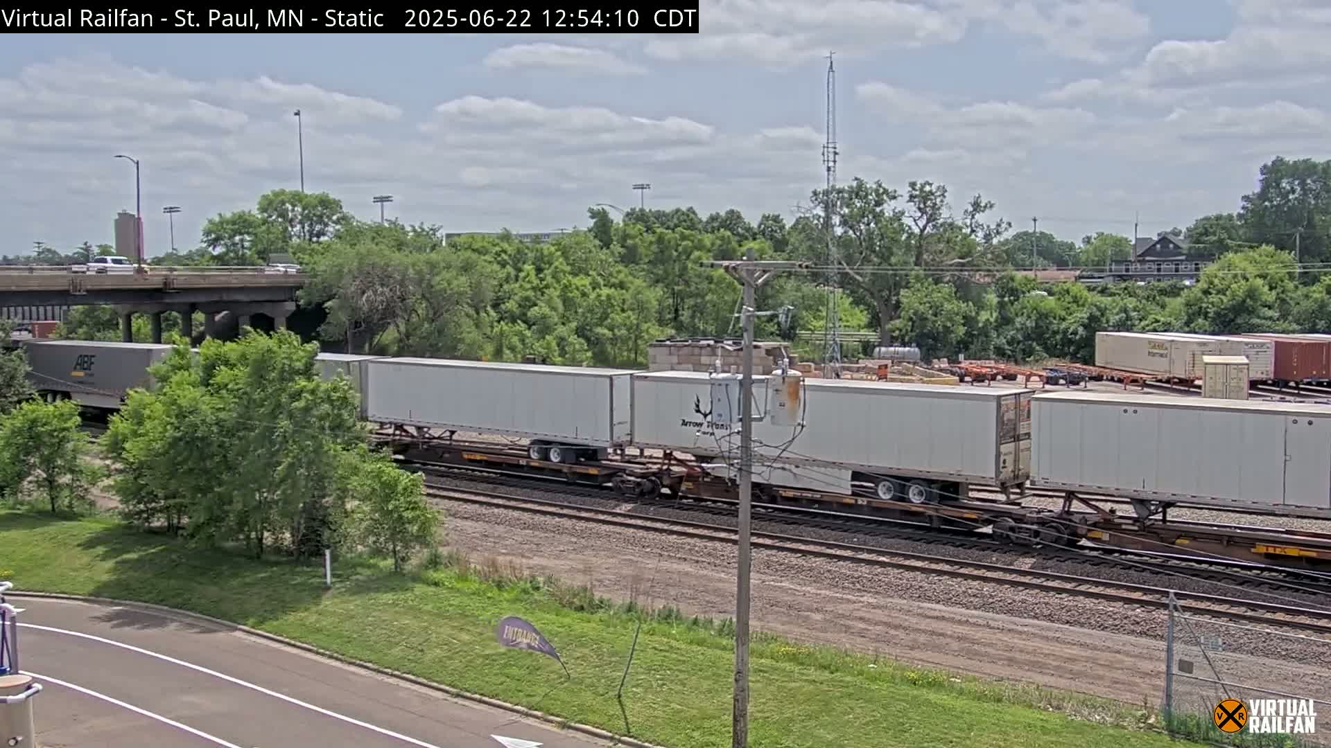 A long freight train with several white shipping containers passes under a bridge on a partly cloudy day.