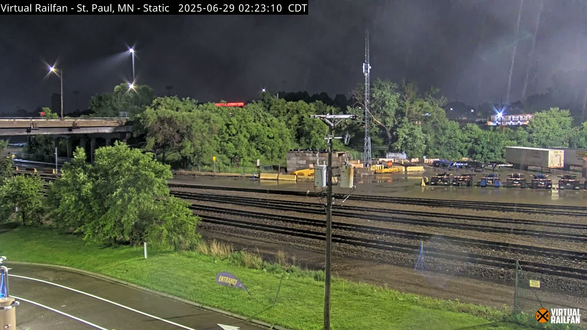 A nighttime view of a flooded rail yard next to a highway overpass under a dark, stormy sky.