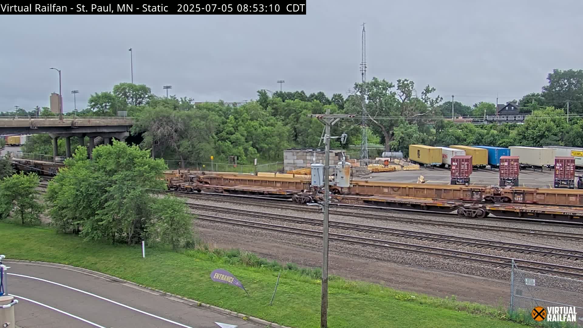 Under an overcast sky, several long freight cars sit on a railway track next to a yard of shipping containers, with a road and overpass visible in the background.