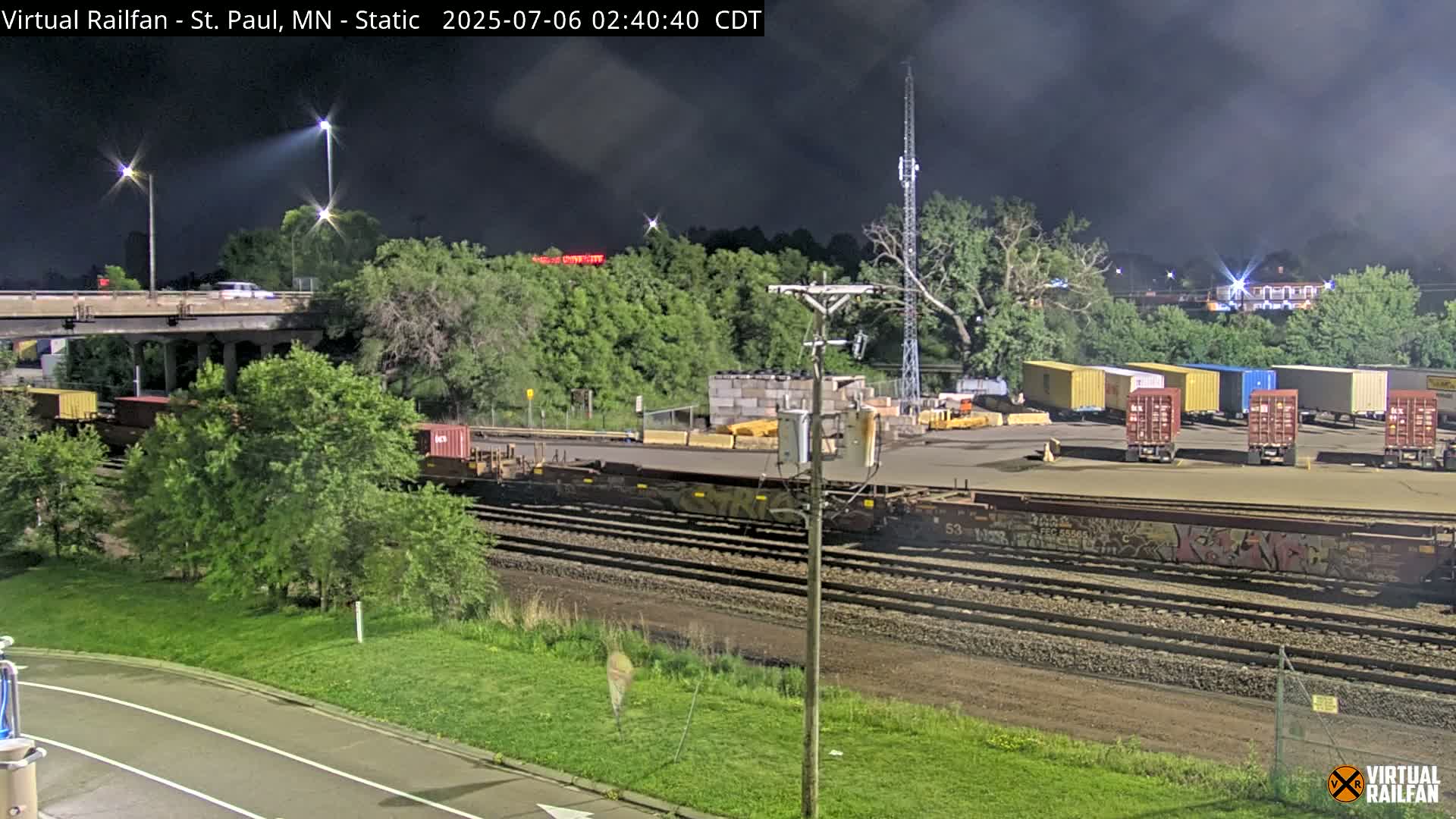 At night, under a dark, cloudy sky, a train sits on tracks near a road and bridge, with several shipping containers visible in a nearby yard.