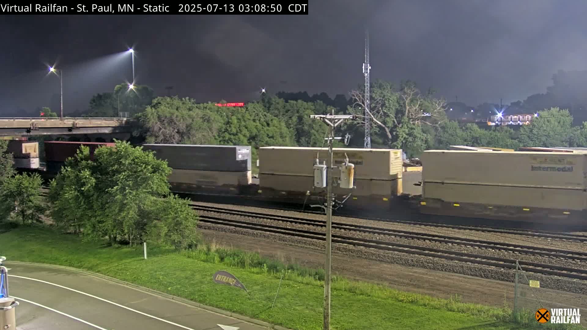 A freight train passes under a bridge at night during overcast conditions.