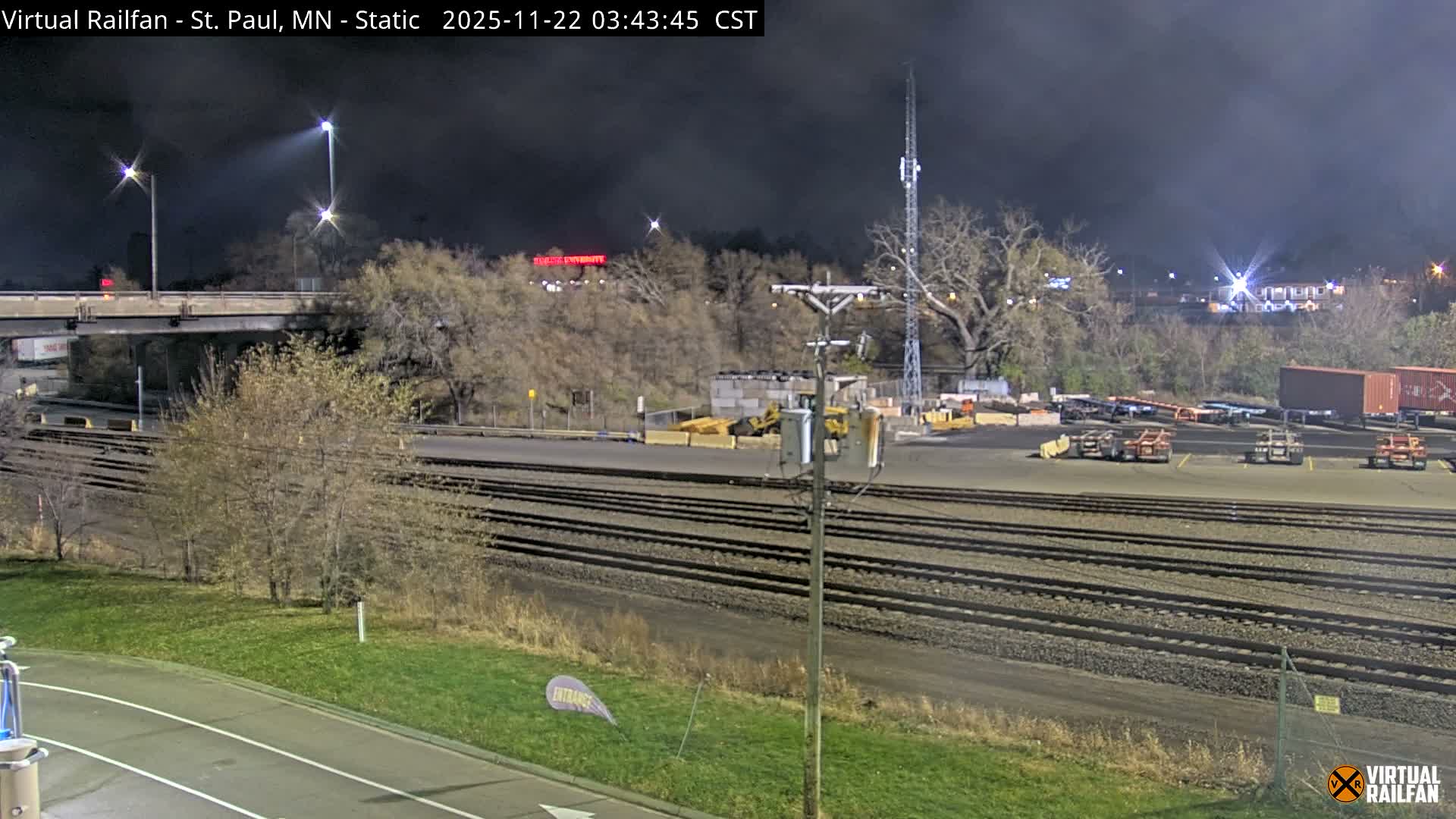 On a clear, dark night, multiple parallel railroad tracks stretch across the foreground, leading to an industrial area filled with heavy machinery, shipping containers, and a tall communication tower, all illuminated by bright streetlights, with a bridge visible overhead on the left.