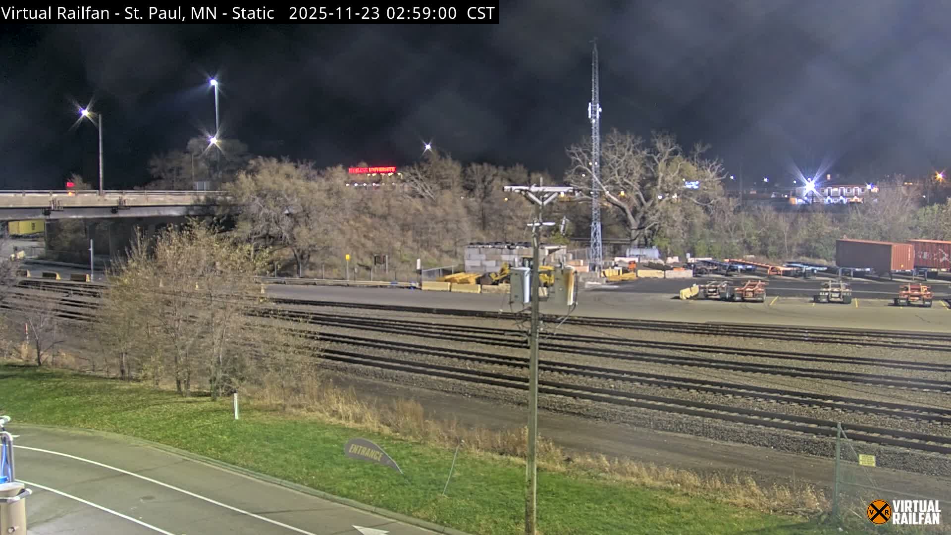 A nighttime scene reveals an extensive rail yard with multiple parallel tracks, shipping containers, and industrial equipment under a dark sky, illuminated by streetlights, with a bridge overhead and distant lights on a hillside.