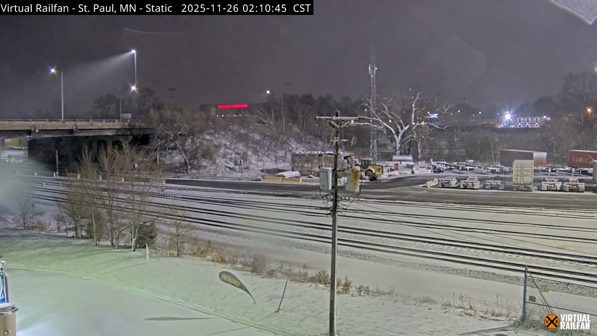 A snow-covered rail yard with numerous parallel tracks stretches across the foreground and midground at night, flanked by bare trees, an elevated bridge, and an industrial storage area with trailers and a bulldozer in the background, all under snowy conditions.