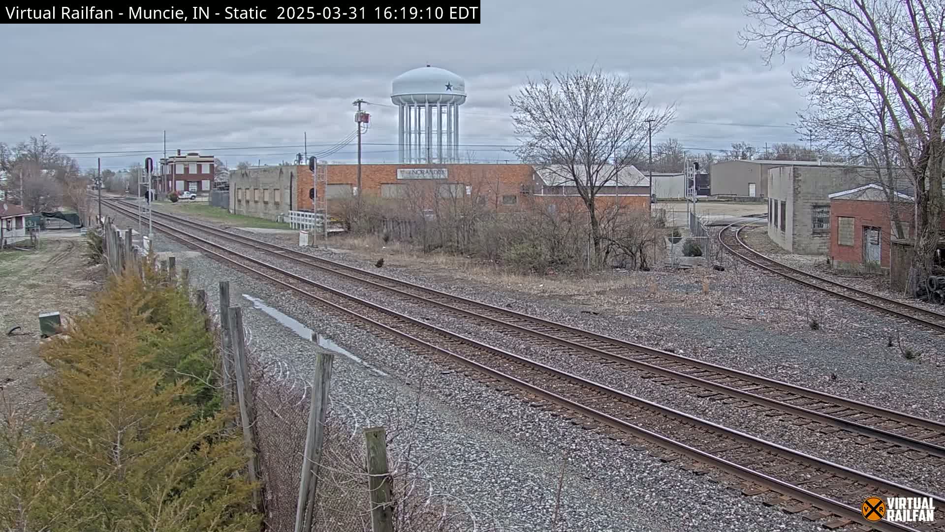 A view of railroad tracks splitting into two directions, with industrial buildings and a water tower in the background under an overcast sky.