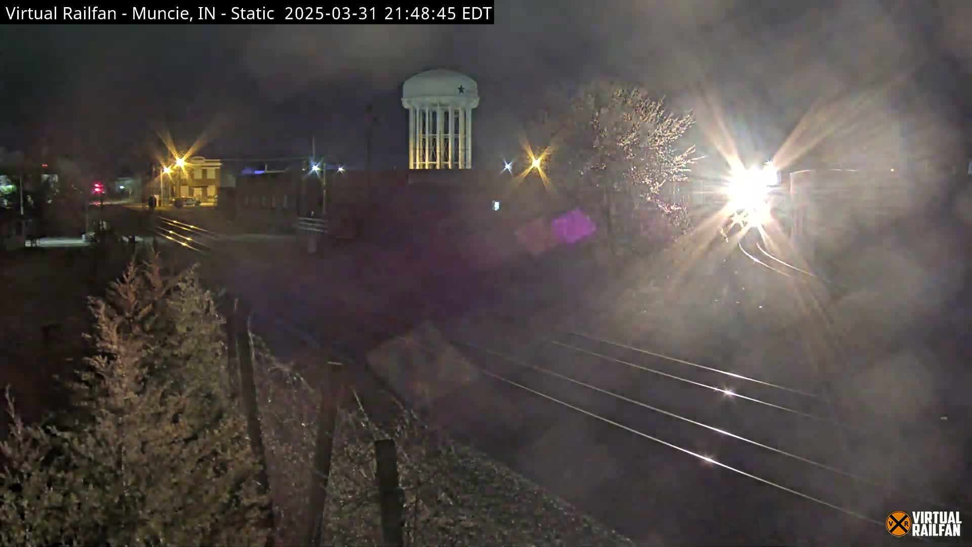 At night, a train approaches a rail crossing near a water tower and some buildings under a dark, clear sky.