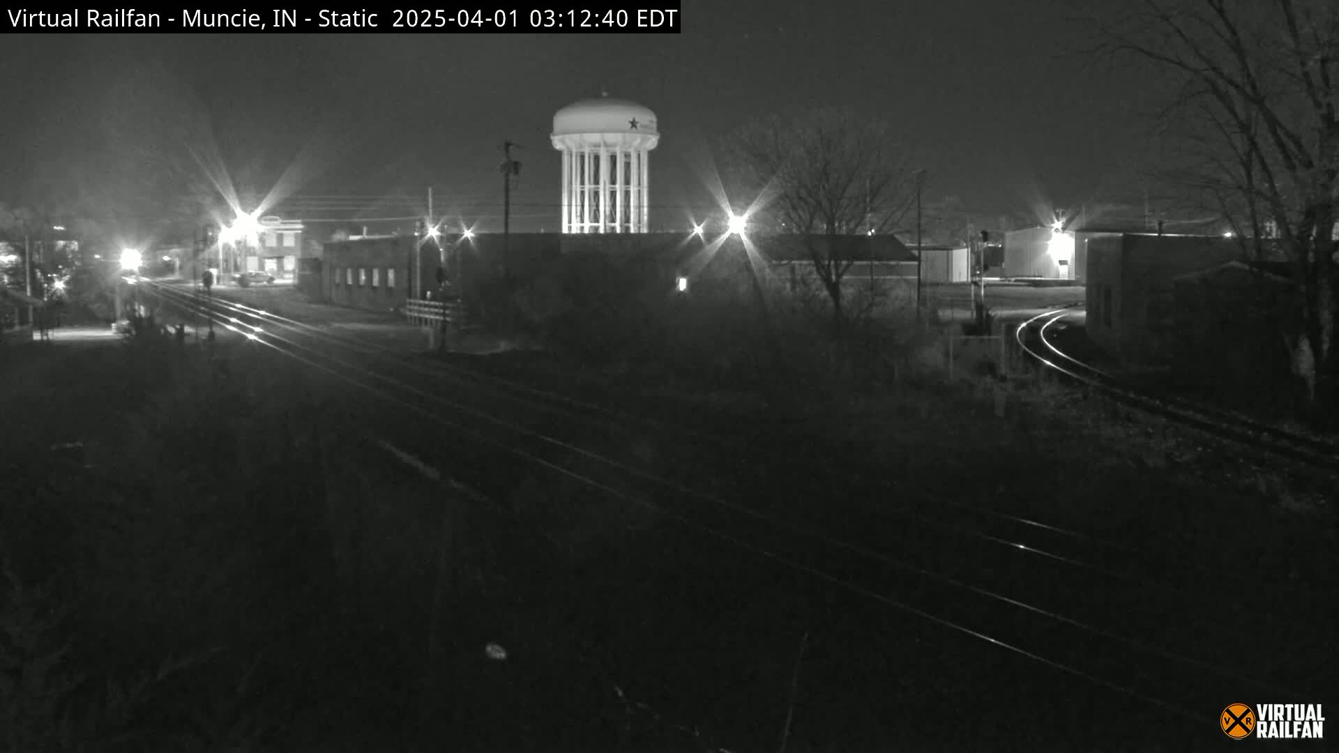 A nighttime view of railroad tracks intersecting a road, with a water tower visible in the background under a clear sky.