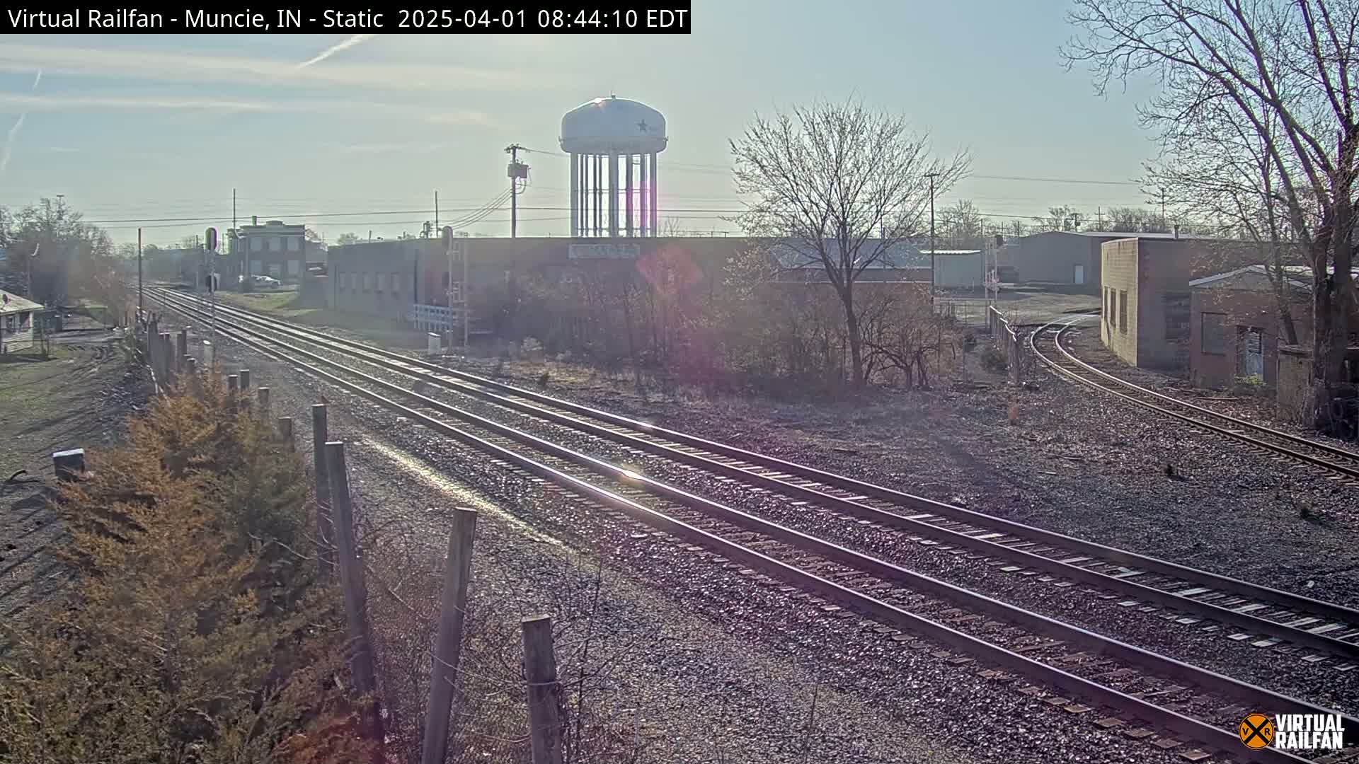 Under a clear, sunny sky, multiple train tracks extend into the distance, passing industrial buildings and a water tower, bordered by bare winter trees and scrubby vegetation.