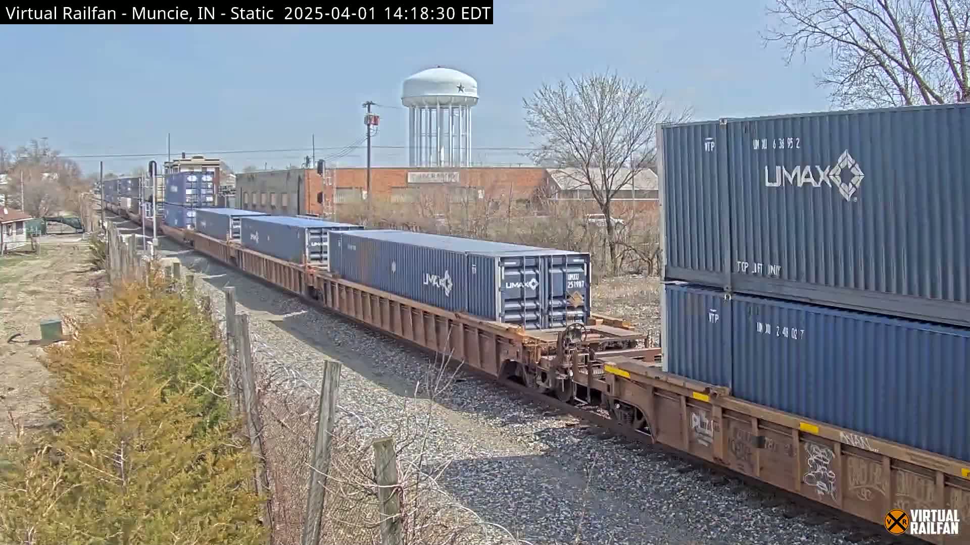 A long freight train carrying multiple shipping containers moves along railroad tracks on a sunny day, passing by industrial buildings and a water tower.